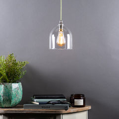 A clear glass pendant light with a frilled design, hanging above a wooden table with a plant, books, and a candle. The room has a neutral grey backdrop.
