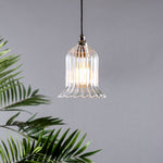 A clear glass pendant light with a fluted design, featuring a metallic finish and suspended by a cable, against a grey background with a green plant to the side.