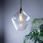 Clear glass diamond shaped pendant light with a visible bulb hanging next to a green plant against a grey background