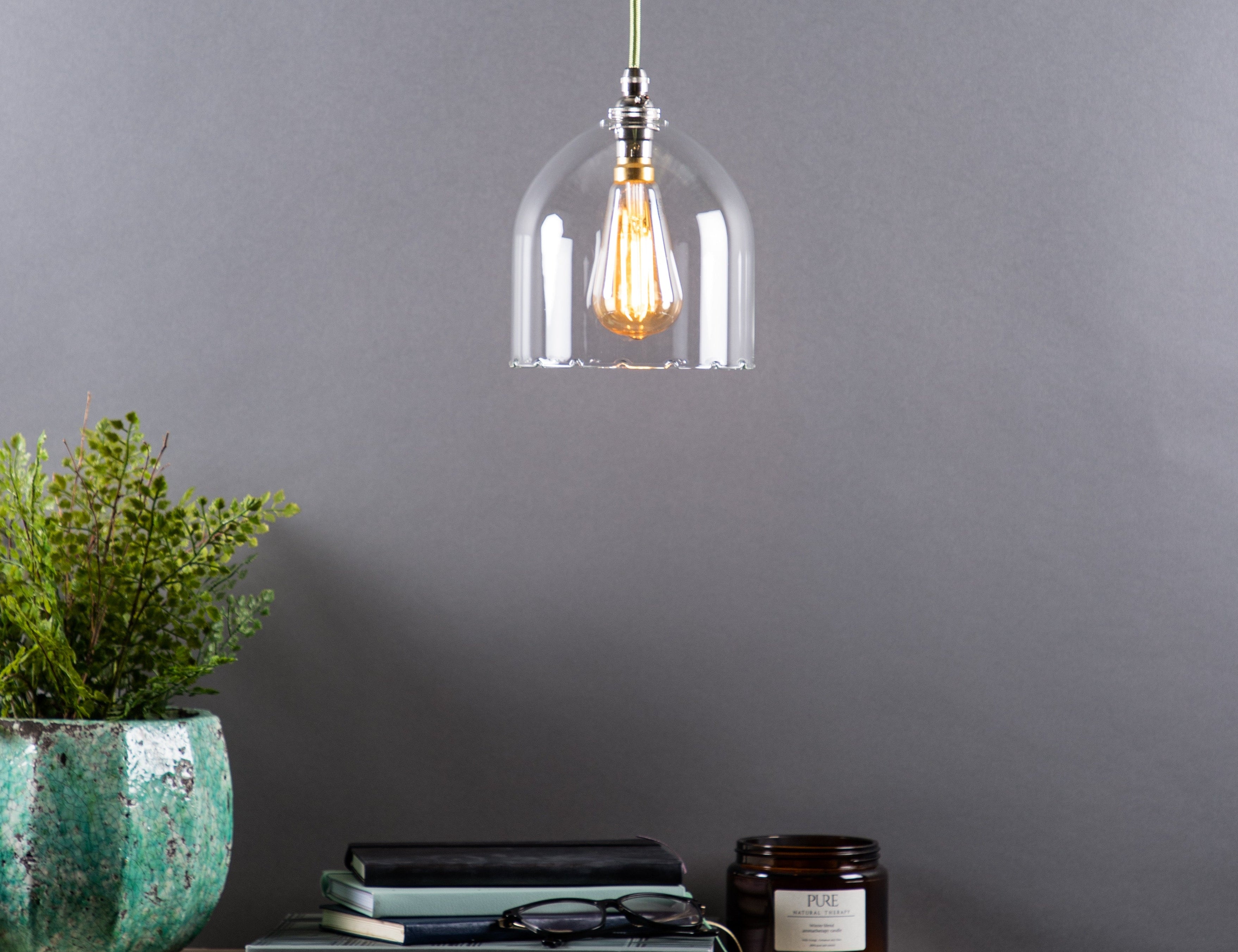 A clear glass pendant light with a frilled design, hanging above a wooden table with a plant, books, and a candle. The room has a neutral grey backdrop.