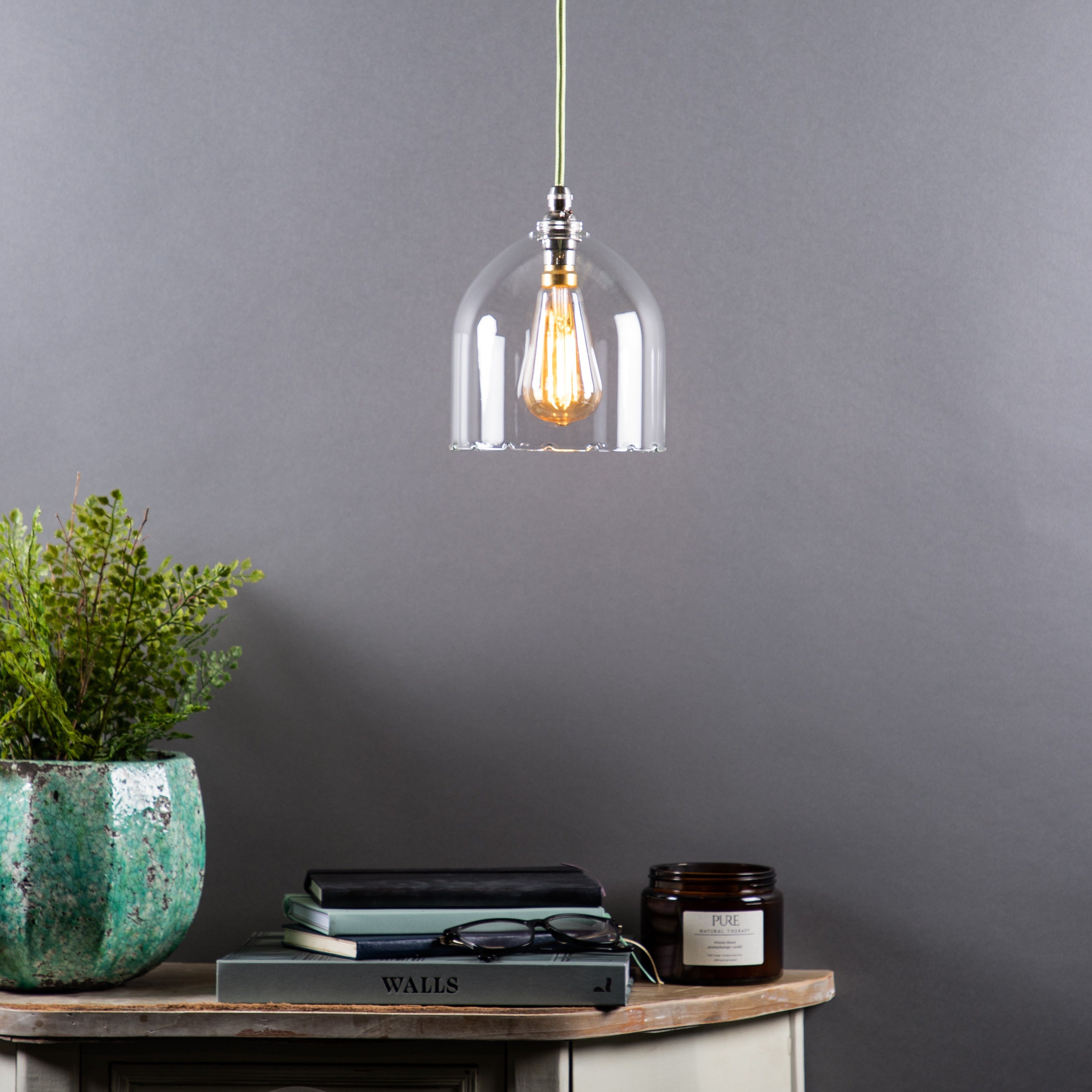 A clear glass pendant light with a frilled design, hanging above a wooden table with a plant, books, and a candle. The room has a neutral grey backdrop.