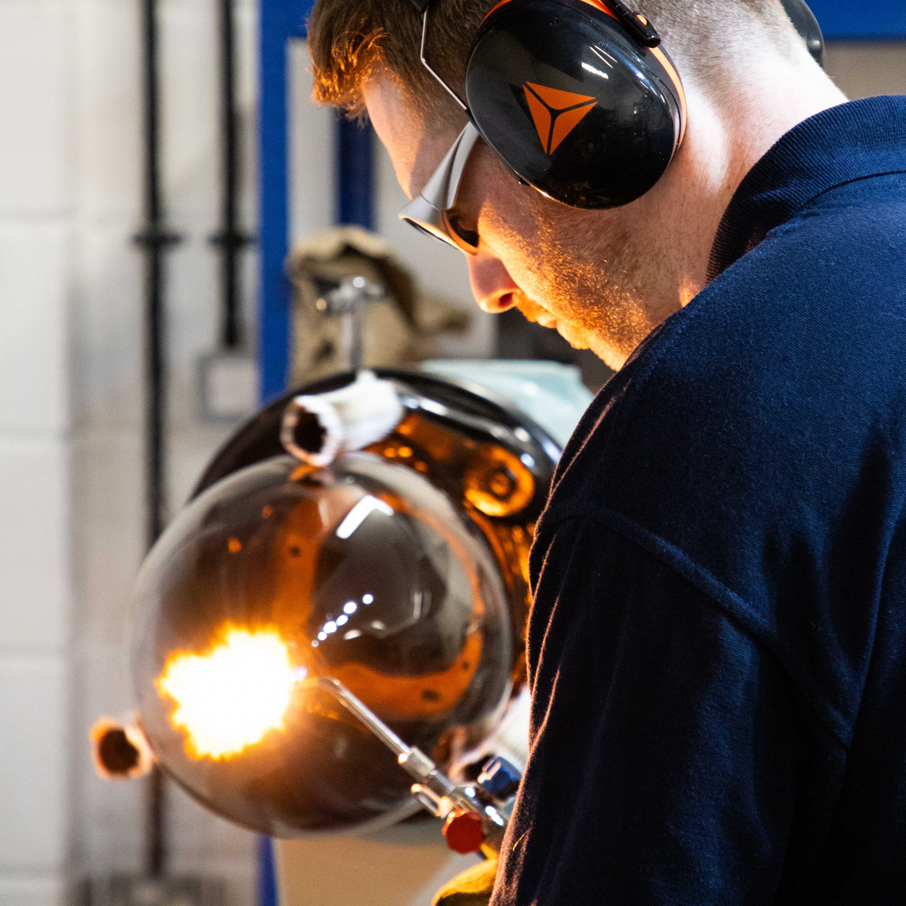 A close-up lifestyle shot of a man wearing protective safety glasses, black over-ear headphones with an orange logo, and a dark navy work jacket, operating a glassblowing furnace in an industrial workshop.
