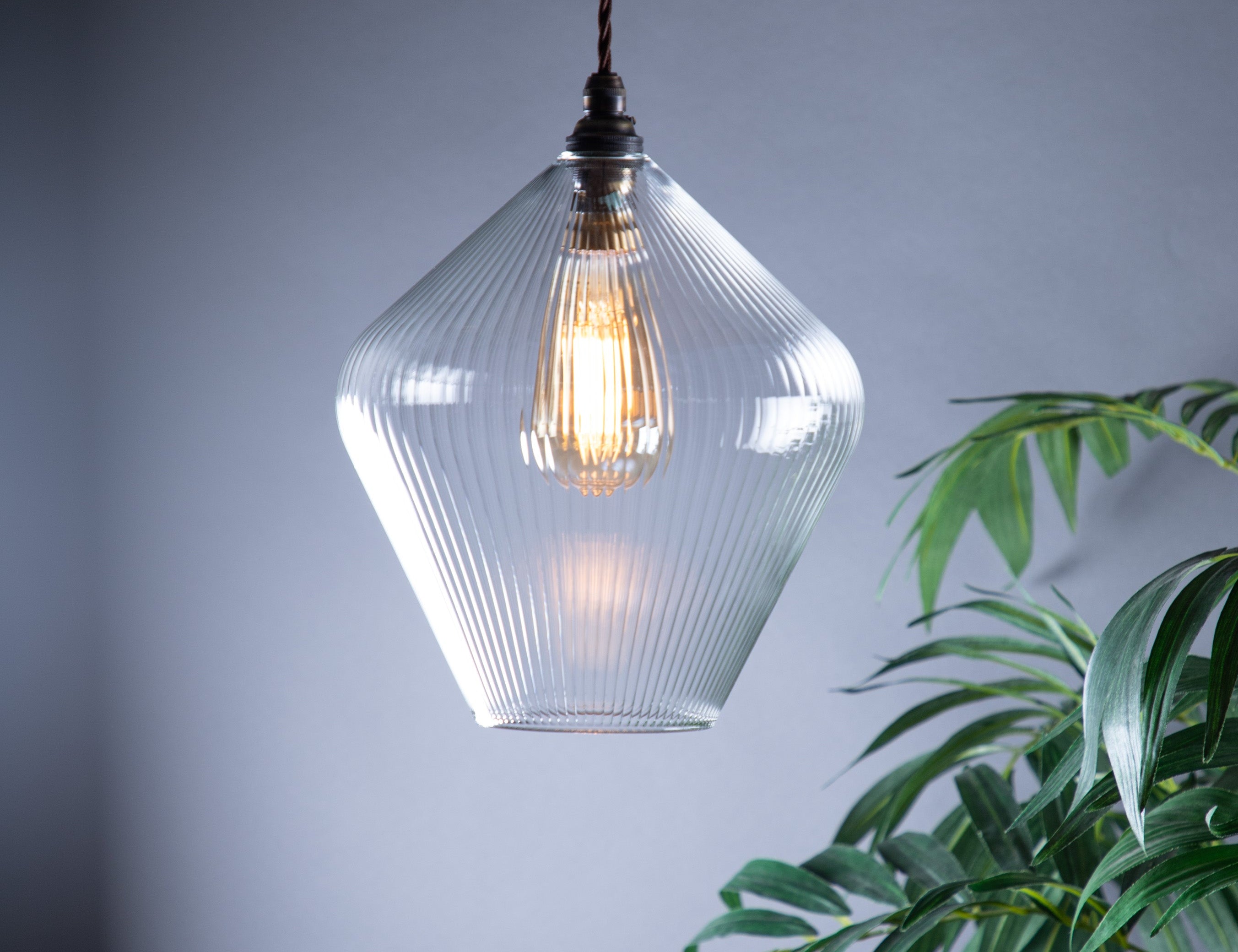 Clear glass diamond shaped pendant light with a visible bulb hanging next to a green plant against a grey background