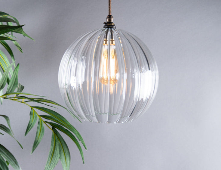 Clear glass pendant light with visible bulb hanging against a grey background with green leaves.