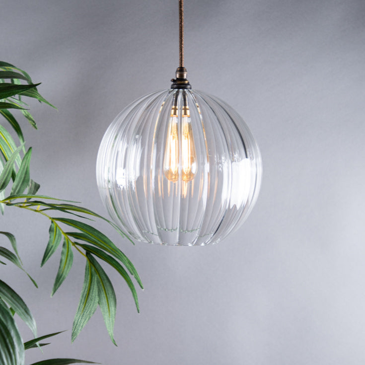 Clear glass pendant light with visible bulb hanging against a grey background with green leaves.